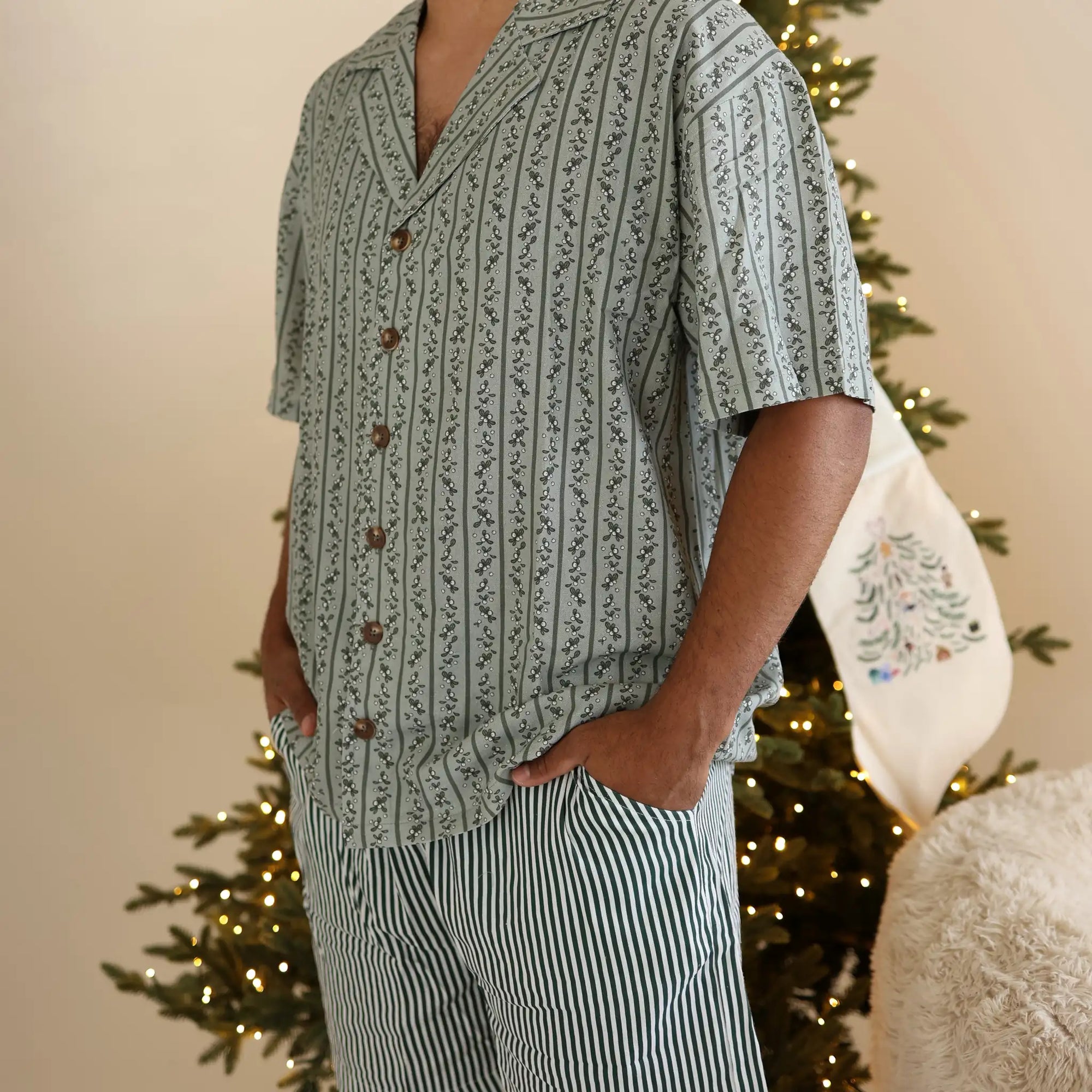 Man in a sage green botanical print shirt and stripe pyjama shorts beside an embroidered Christmas stocking