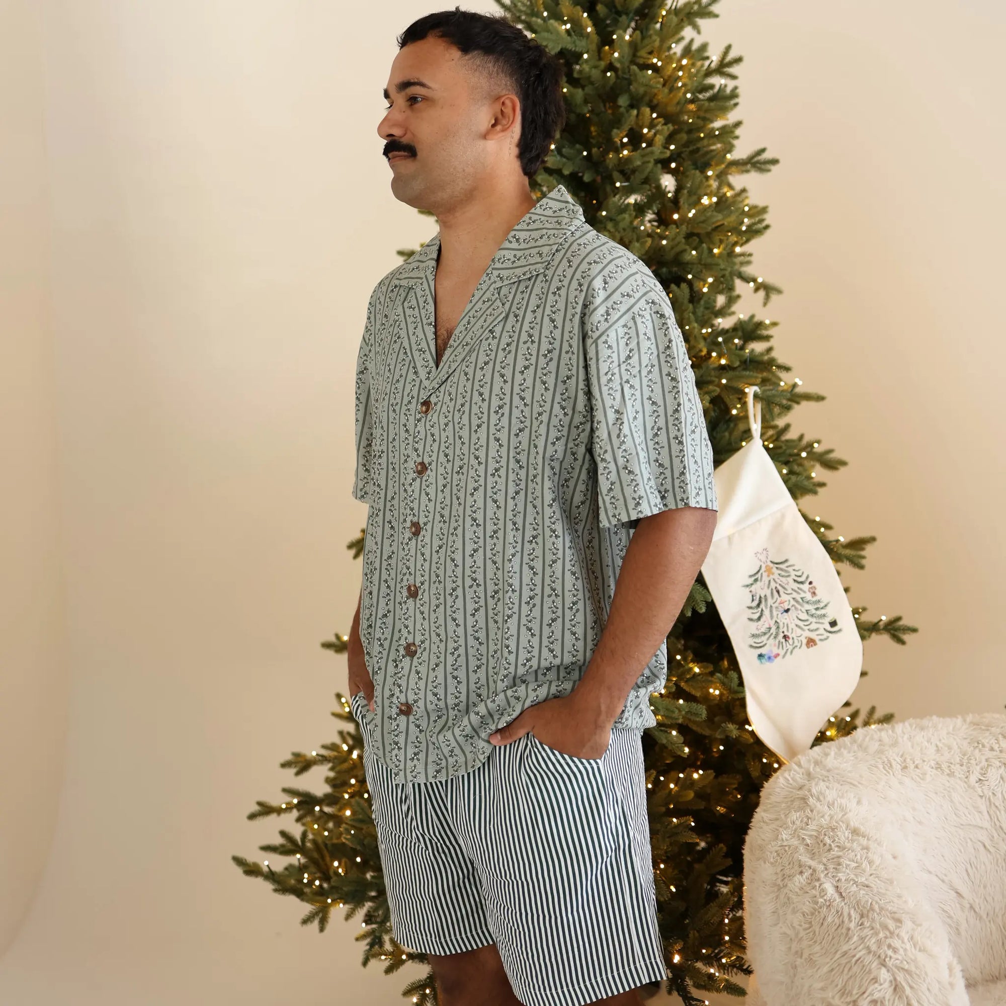 Man in a sage green botanical print short sleeve shirt and stripe shorts pyjama set by a lit Christmas tree
