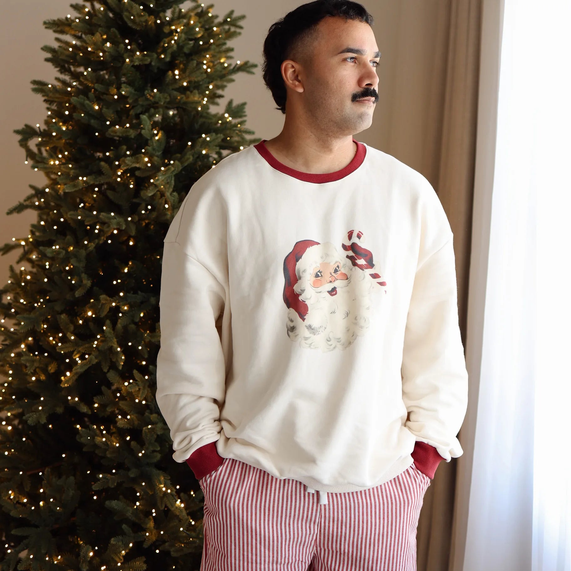 Man in a cream vintage Santa print sweatshirt with red trim and stripe pyjama pants by a lit Christmas tree