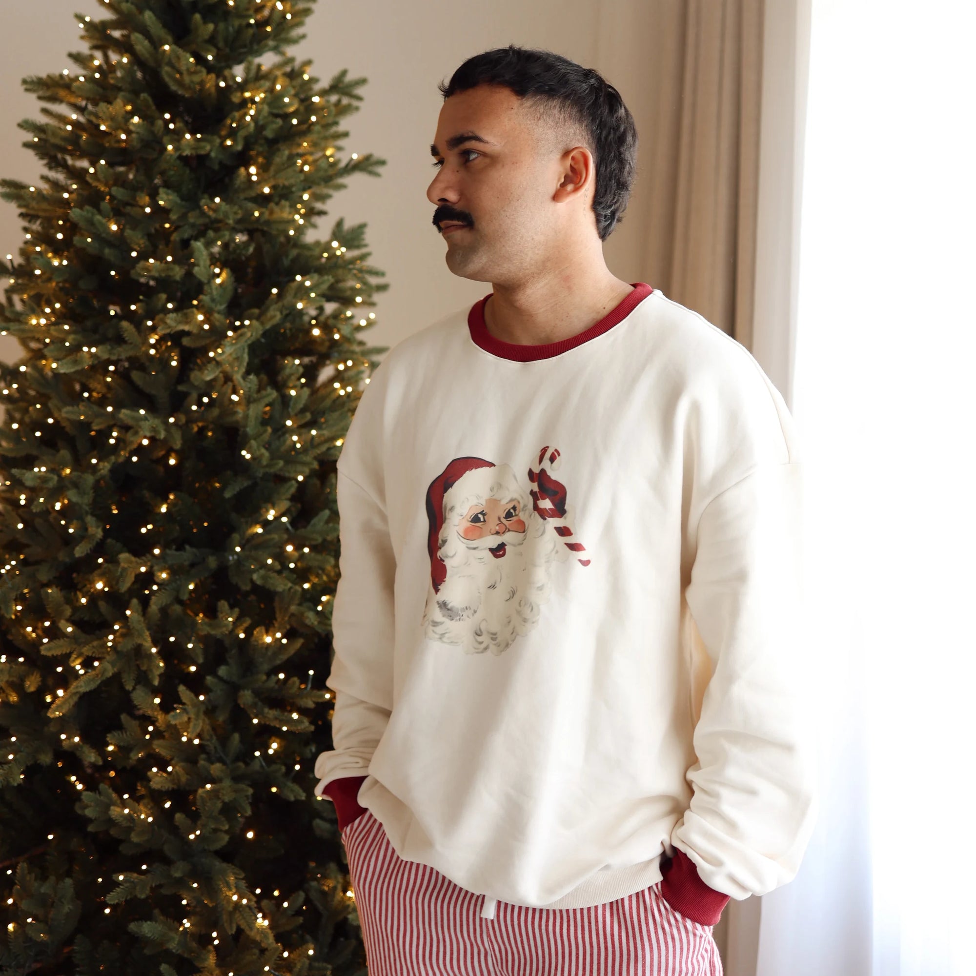 Man in a vintage Santa print sweatshirt and red stripe pyjama pants beside a lit Christmas tree