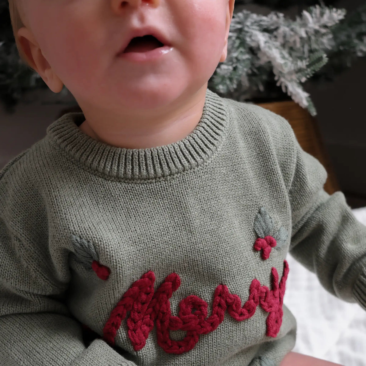A close-up of a baby wearing an olive green knit sweater with red 'Merry' lettering and holly accents on the front, set against a soft holiday background.