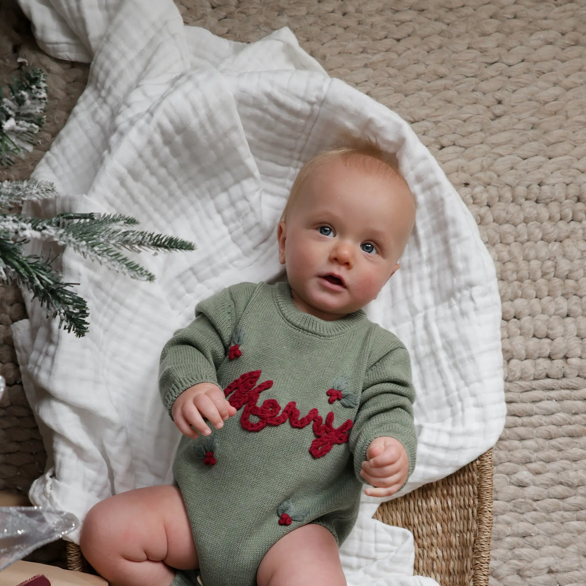 A baby wearing a sage green knit sweater with 'Merry' embroidered in red across the chest, sitting on a white muslin blanket on a beige textured rug.
