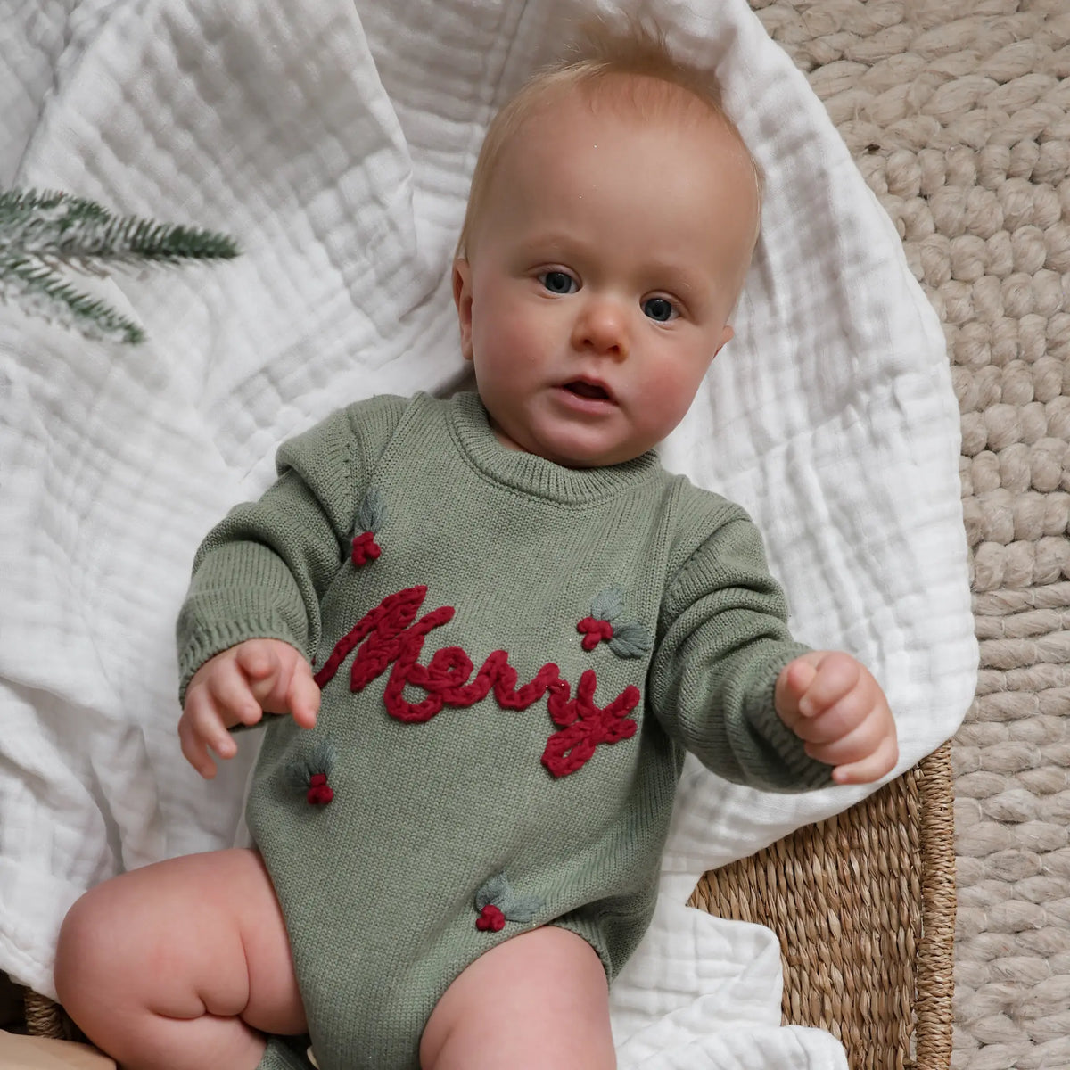 A baby wearing an olive green knit romper with red 'Merry' lettering and small red and gray holly accents, sitting on a white textured blanket.