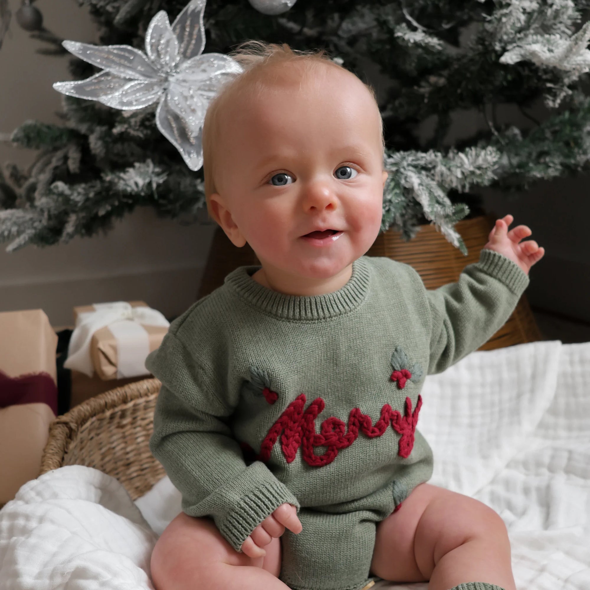 A baby sitting in a wicker basket wearing an olive green knit sweater with red 'Merry' lettering and holly details, posed in front of a decorated Christmas tree.