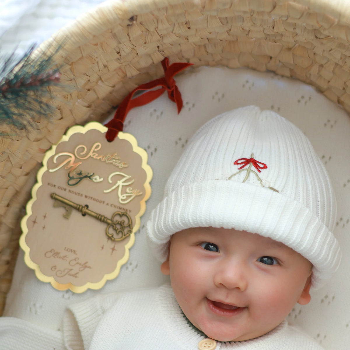 A baby wearing a white ribbed knit beanie with a small red bow and gold key embroidery, sitting in a woven basket next to a hanging gift tag that reads 'Santa's Magic Key'.