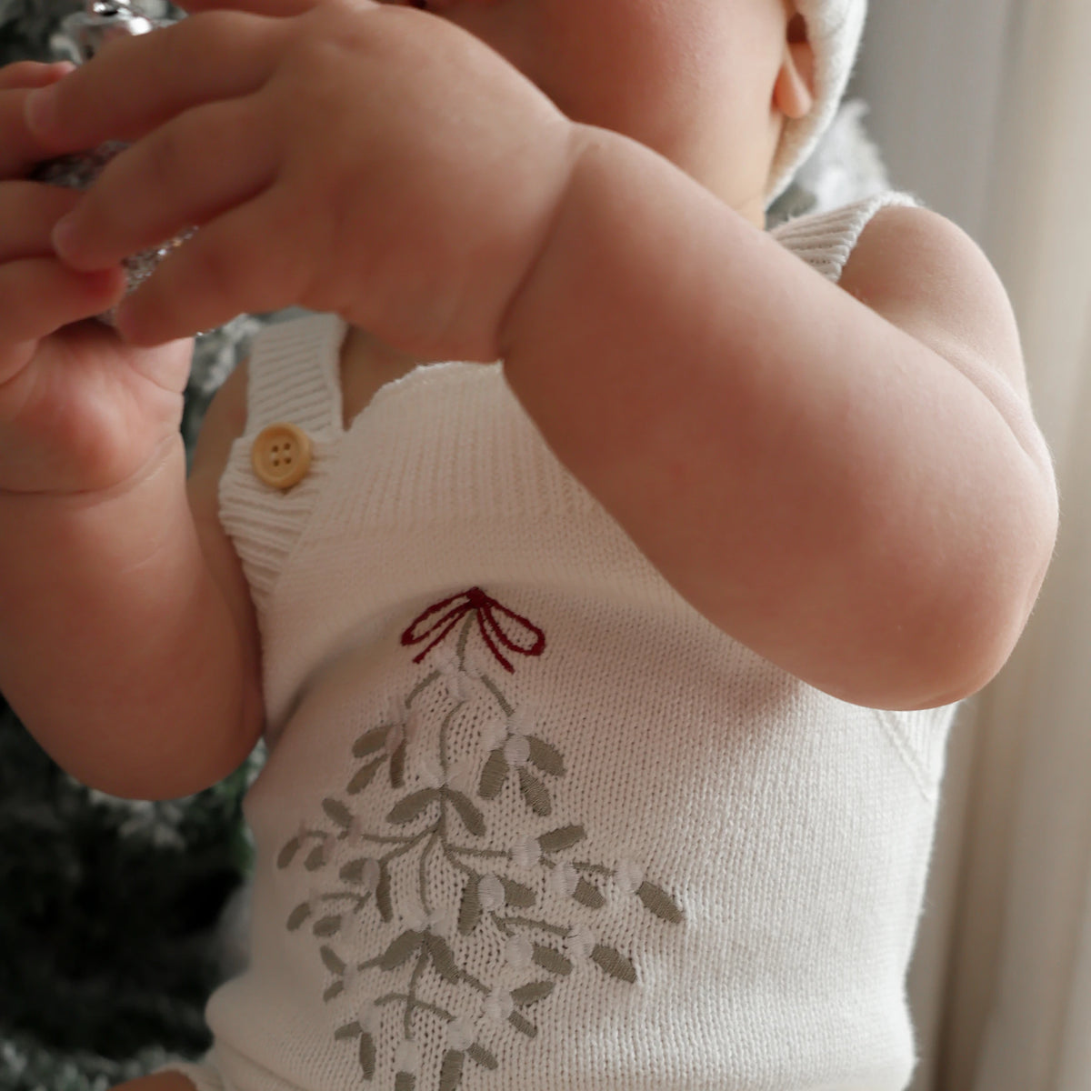 A close-up of a baby wearing a cream knit sleeveless bodysuit with a gray leafy design and a small red bow motif on the front, featuring a wooden button closure.