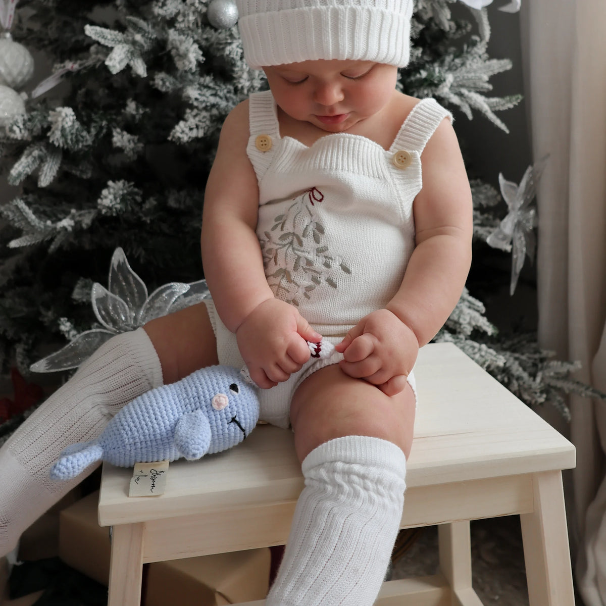 A baby wearing a white knit sweater vest with a floral print, a matching white knit beanie, and white ribbed socks, sitting on a small wooden stool in front of a decorated Christmas tree. The baby is holding a small blue knitted animal toy.
