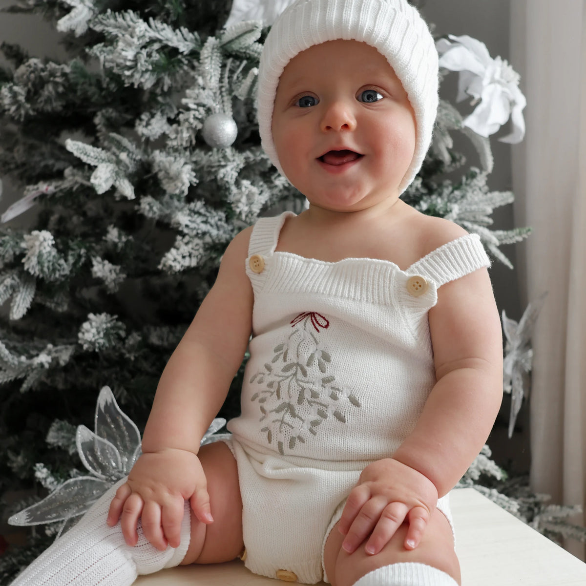 A baby wearing a white knit bonnet, white knit overalls with a gray embroidered mistletoe design, and white knit socks, sitting in front of a decorated Christmas tree.