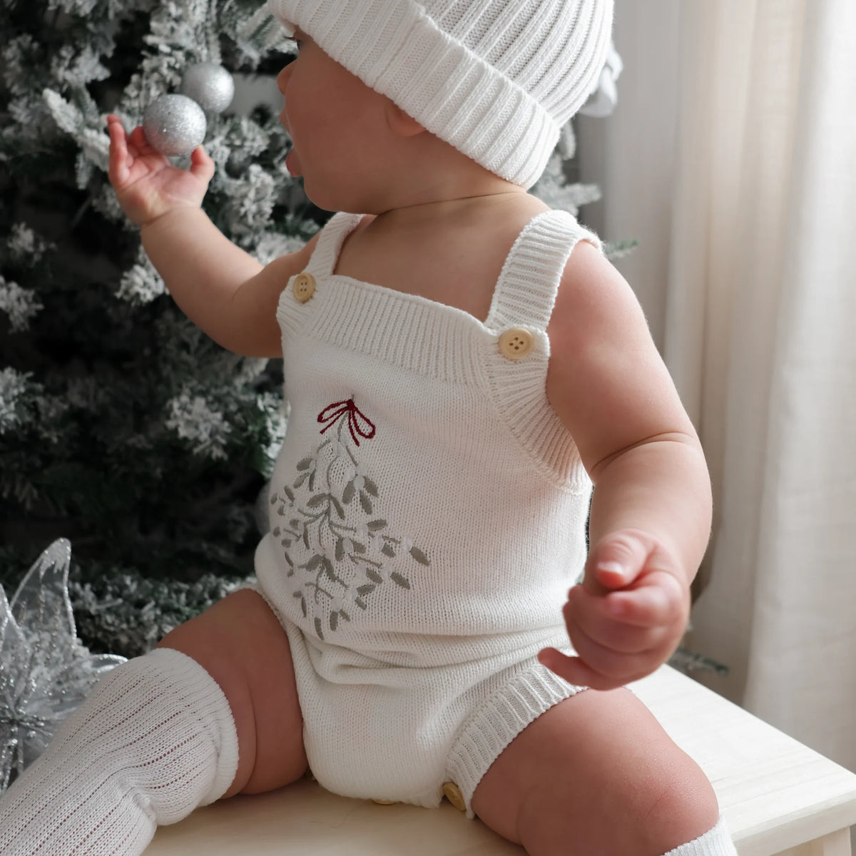 A baby wearing a cream knit romper with a gray mistletoe design and a red bow, matching knit hat, and leg warmers, sitting next to a decorated Christmas tree.