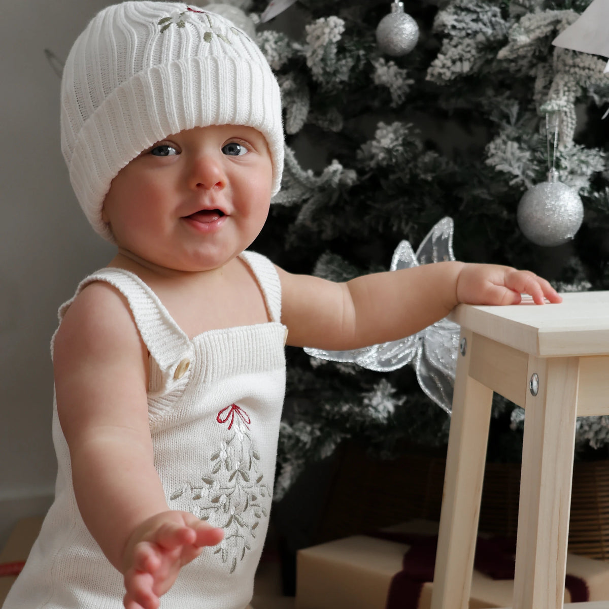 A baby wearing a white ribbed knit beanie and matching white sleeveless romper with embroidered floral details, standing beside a light wood stool in front of a decorated Christmas tree.