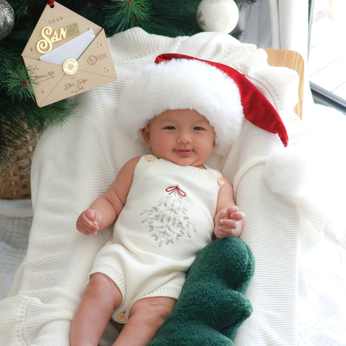 A baby lying on a white blanket, wearing a red Santa hat and a cream holiday romper with a white tree design. A green knit stocking is at the baby's side, and a Christmas-themed envelope prop is near the top left.