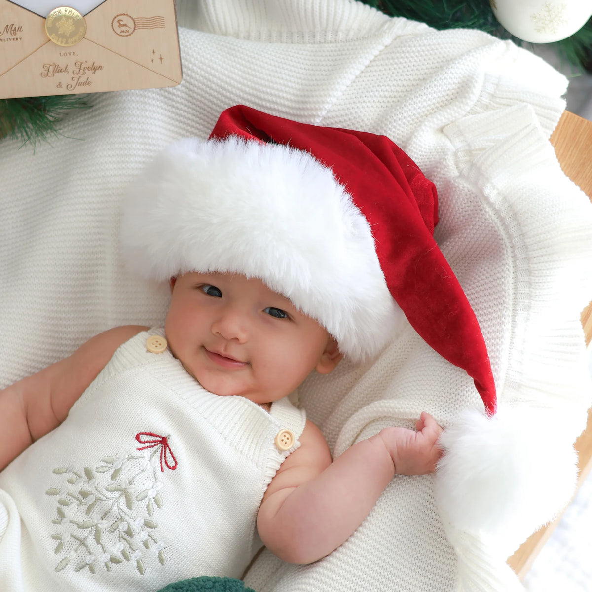 A baby wearing a red Santa hat with white faux fur trim and a white knit holiday romper, lying on a white knit blanket.