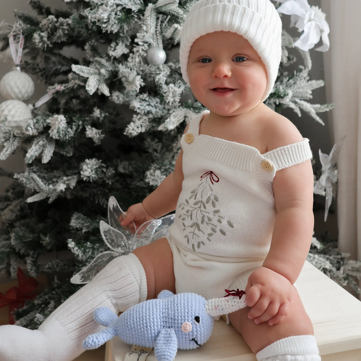 A baby wearing a white knit beanie, white off-the-shoulder knit romper with a leaf embroidery, and white knit booties, sitting in front of a decorated Christmas tree and holding a blue crocheted toy.
