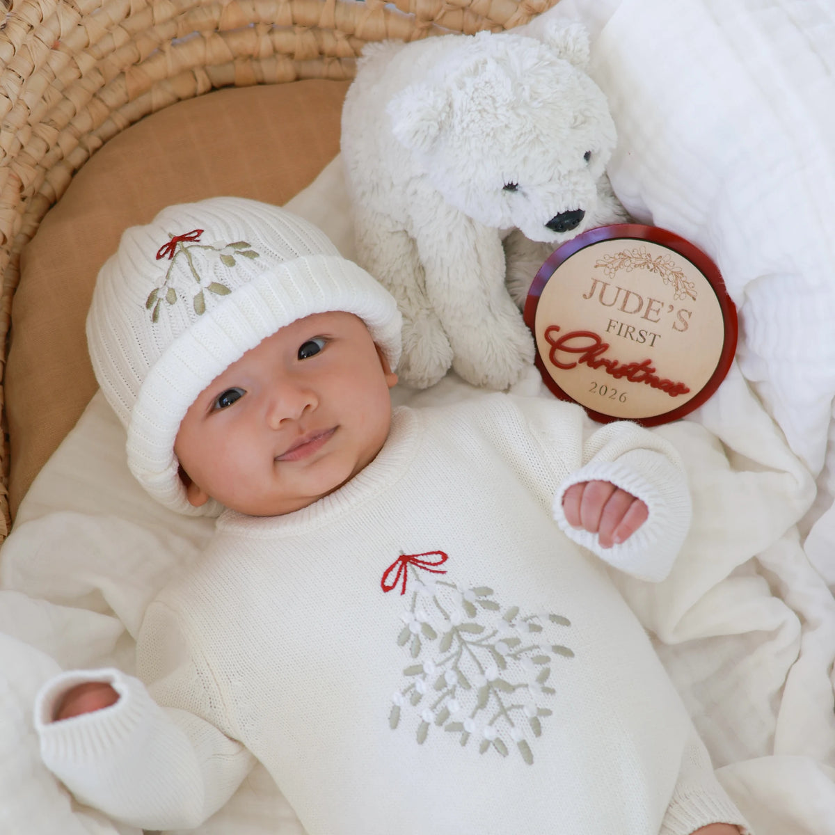 A baby wearing a cream knit hat and matching long-sleeve romper with a gray mistletoe design and small red bow, lying in a woven basket next to a white teddy bear and a round wooden keepsake plaque.