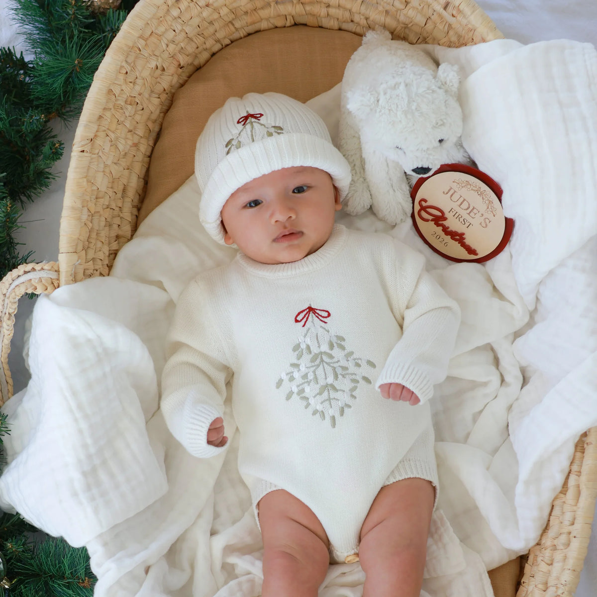 Baby in a white mistletoe knit sweater romper and matching beanie, lying in a woven bassinet with a plush bear and a personalised Jude's First Christmas 2026 keepsake disc