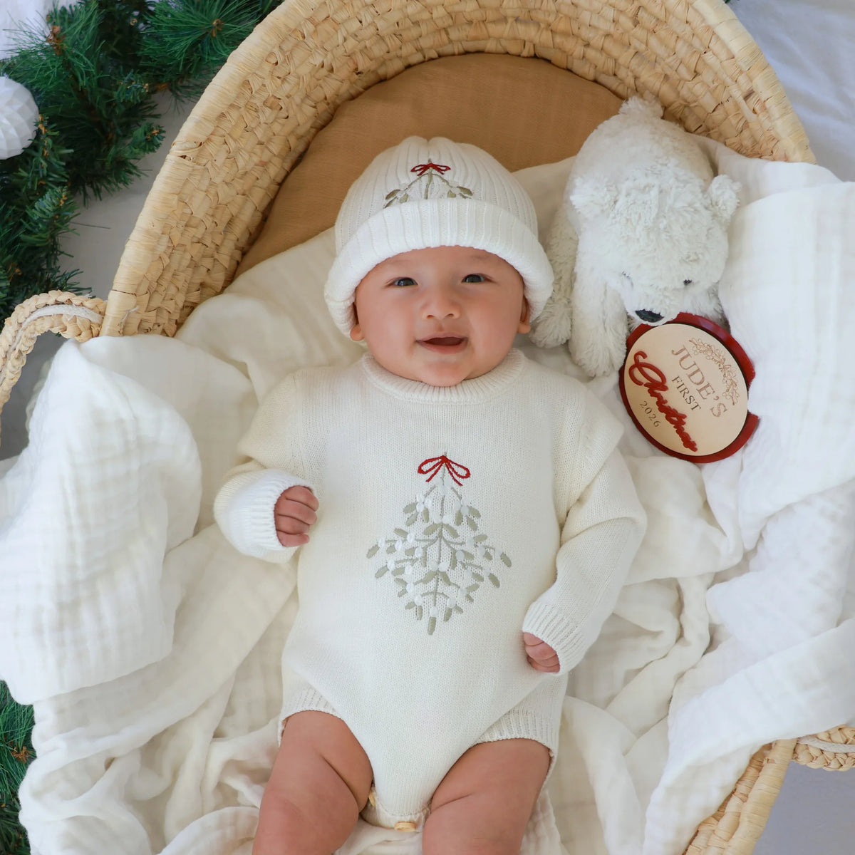 A baby wearing a cream knit bodysuit and matching hat with a gray mistletoe design and red bow, lying in a woven basket with a white plush toy and white muslin blankets.