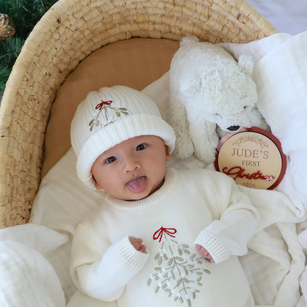 Smiling baby in a white mistletoe knit sweater romper and matching beanie, lying in a woven bassinet with a plush bear and a Jude's First Christmas 2026 keepsake disc