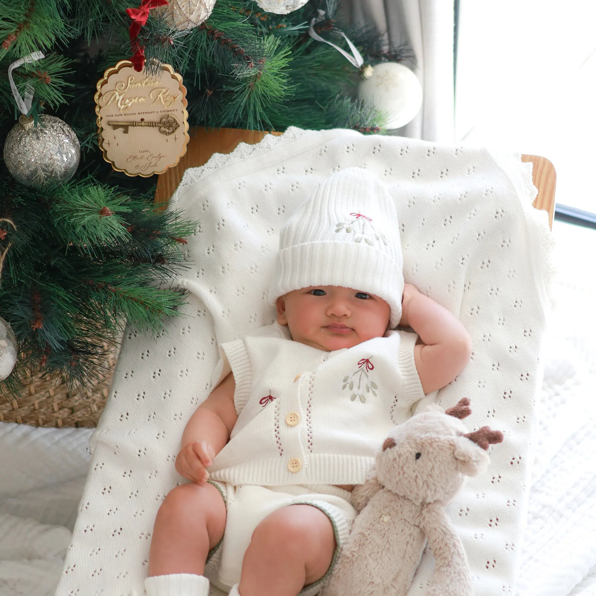 A baby wearing a cream knit hat and matching cardigan lies on a white textured blanket next to a Christmas tree and a stuffed toy, with a window light shining in from the side.