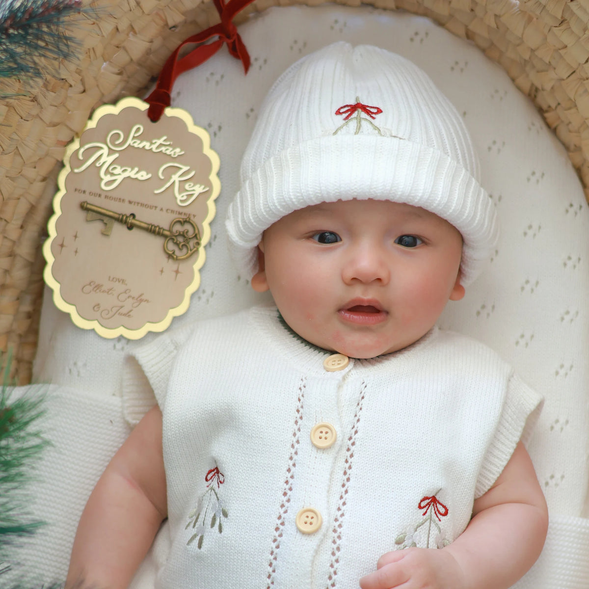 A baby wearing a white knit Santa hat and matching white knit vest with embroidered red bows and green leaves, posed in a woven basket. A hanging tag reads 'Santa's Magic Key' with a decorative key illustration.