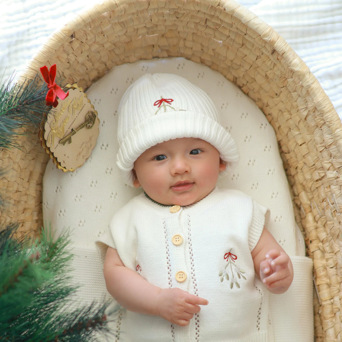 Baby in a white mistletoe knit vest and shorts set with a matching embroidered beanie, lying in a woven bassinet beside a Santa's Magic Key ornament