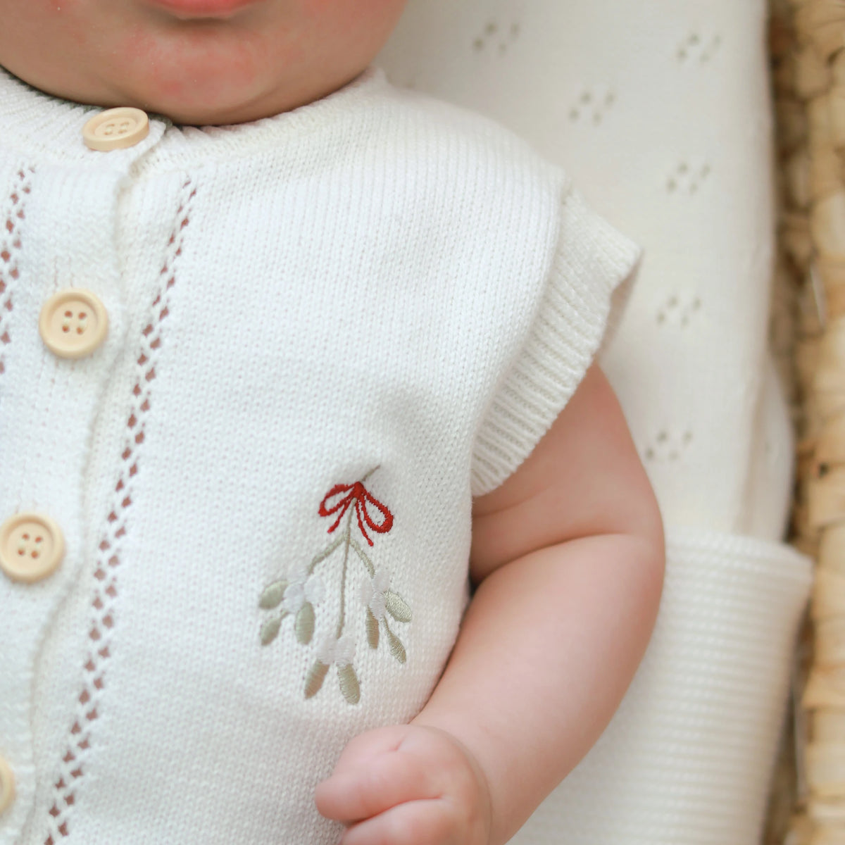 Close-up of a baby wearing a cream knit cardigan with wooden buttons and a small embroidered mistletoe design on the chest.
