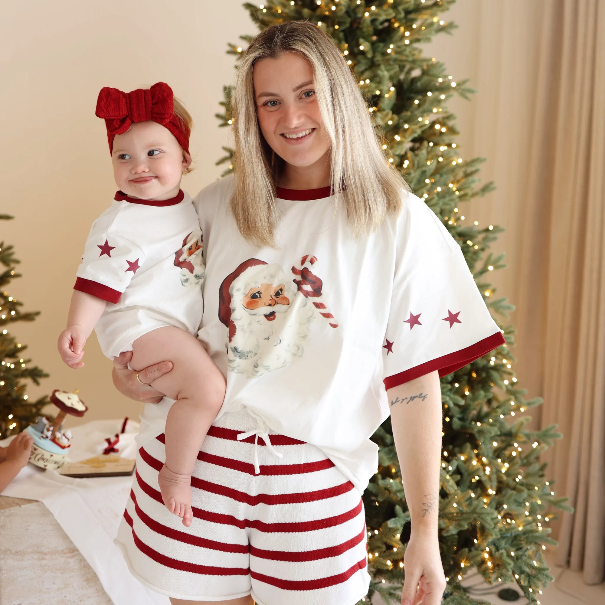 Smiling mum holding a baby in matching vintage Santa print tees and red stripe shorts beside a lit Christmas tree