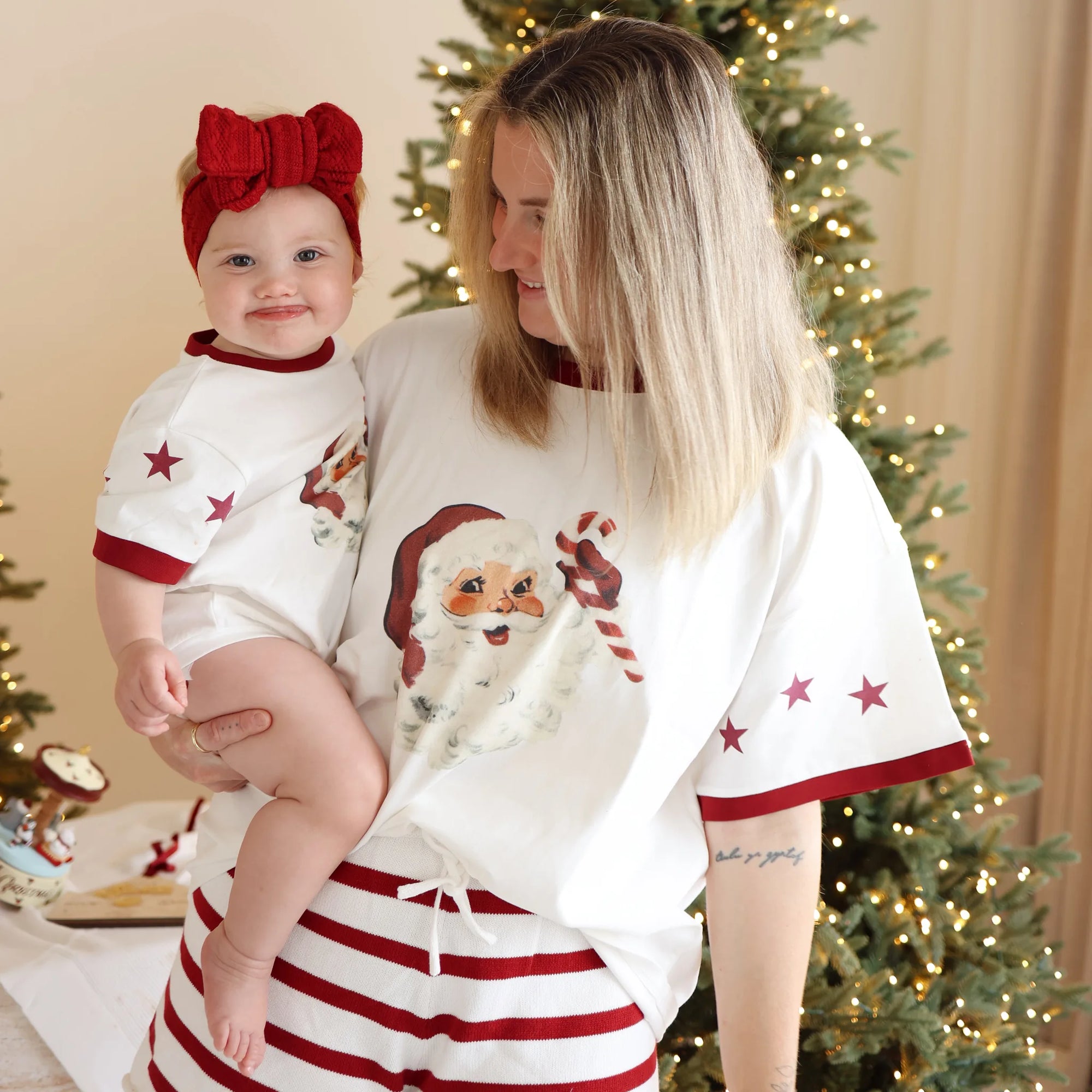 Mum holding a baby in a red knit bow headband, both in matching vintage Santa print tee pyjama sets
