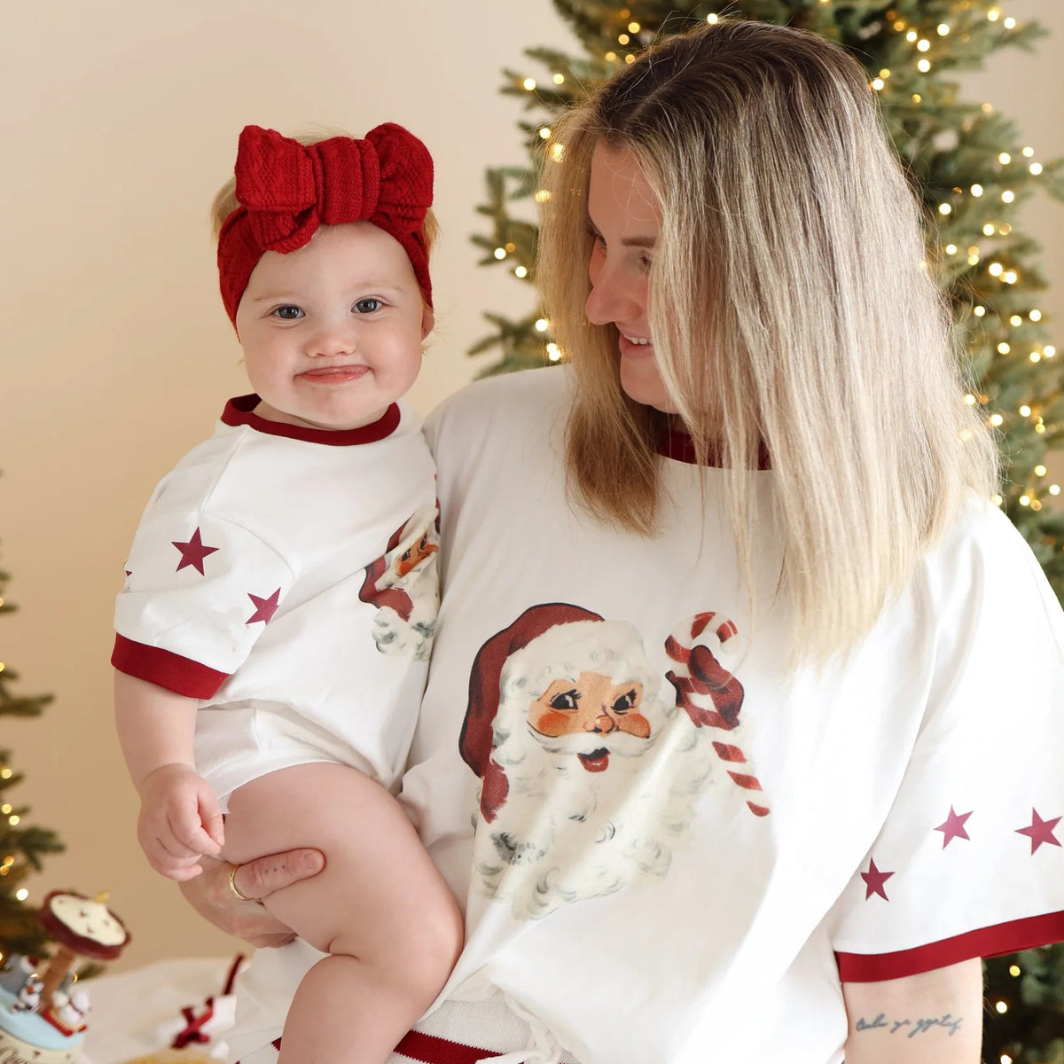 Mum and baby in matching vintage Santa print tees, baby wearing a red knit bow headband, in front of a lit Christmas tree