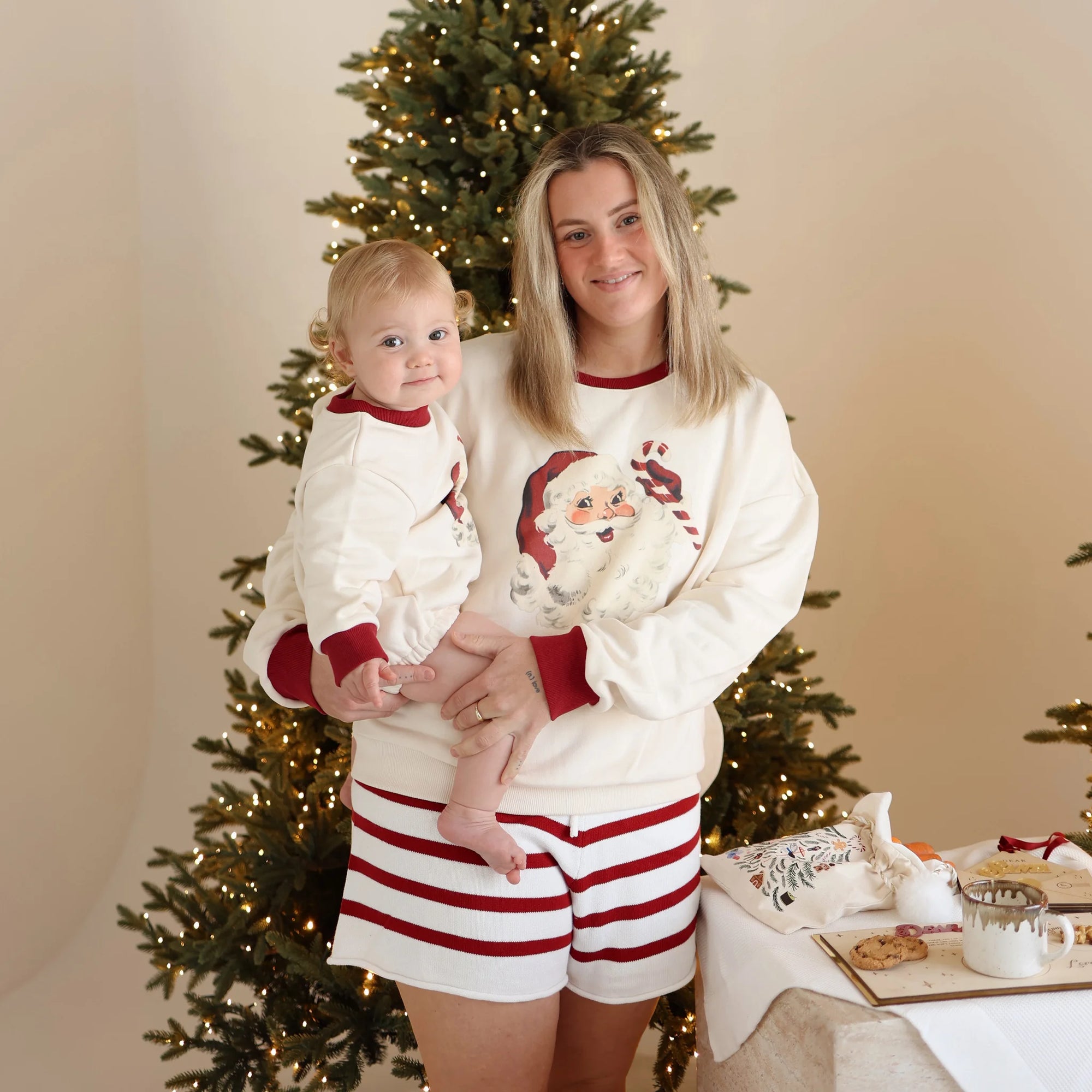 Mum holding a baby in matching vintage Santa print jumpers and red stripe knit shorts beside a lit Christmas tree