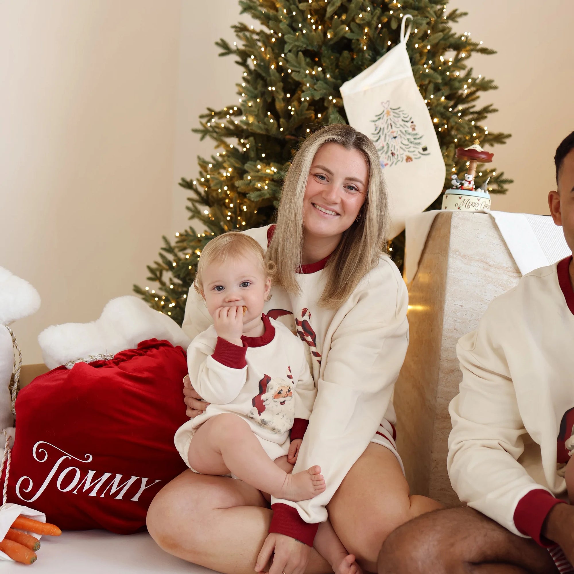 Mum holding a baby, both in vintage Santa print jumpers, beside a personalised red velvet Santa sack embroidered with the name Tommy