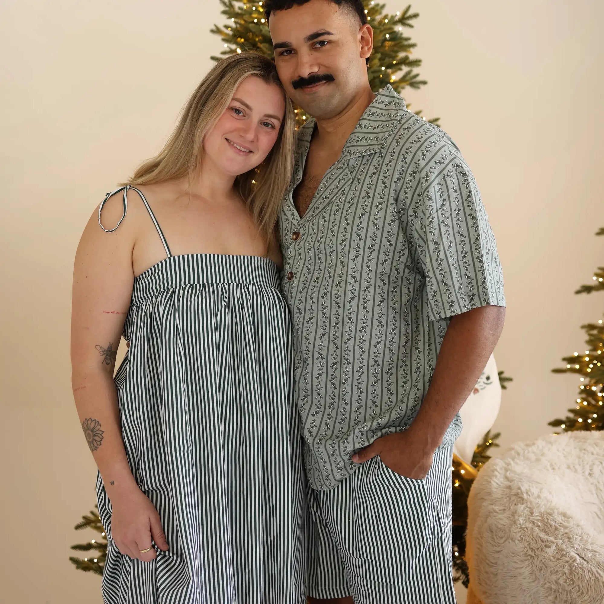 Couple in matching green botanical print and stripe Christmas pyjamas beside a lit Christmas tree