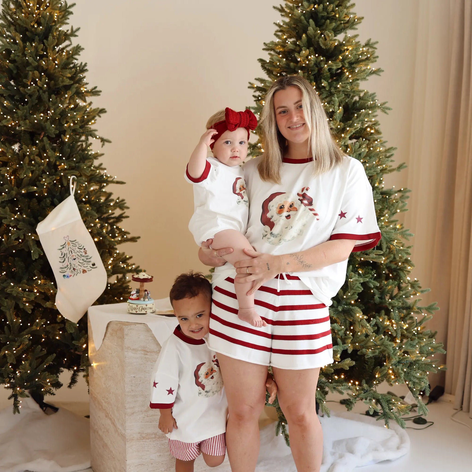 Mum and two children in matching vintage Santa print pyjamas with red stripe shorts, in front of lit Christmas trees