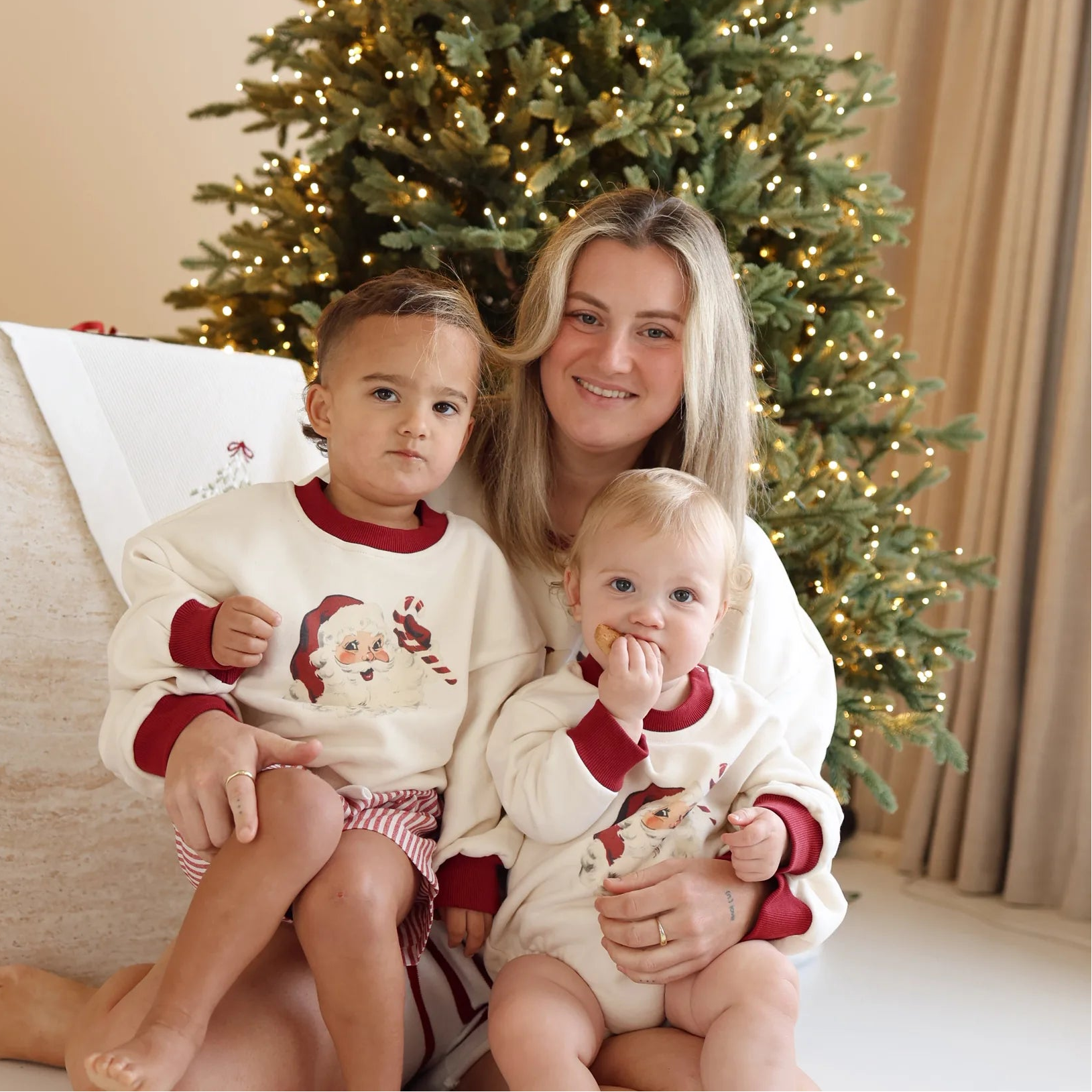 Mum holding two children in matching vintage Santa print jumpers in front of a lit Christmas tree