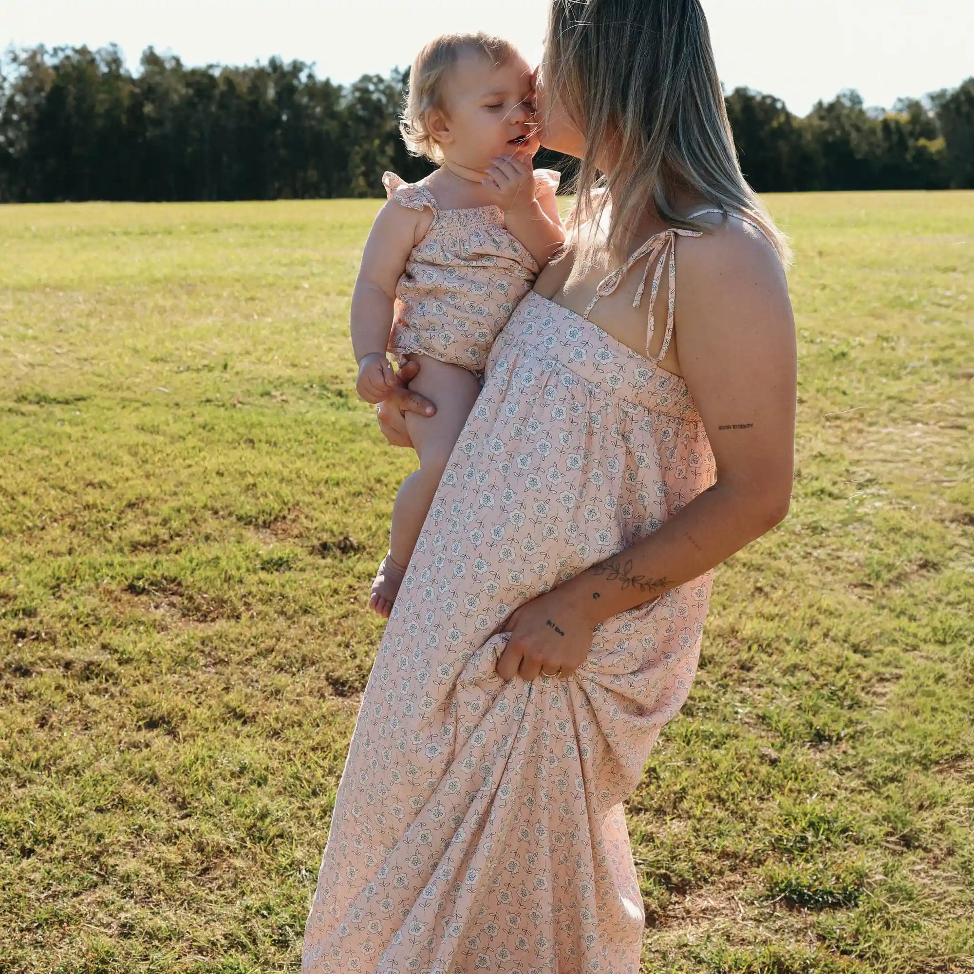 Mum in a floral print maxi dress kissing her baby, who wears a matching floral romper, in a field