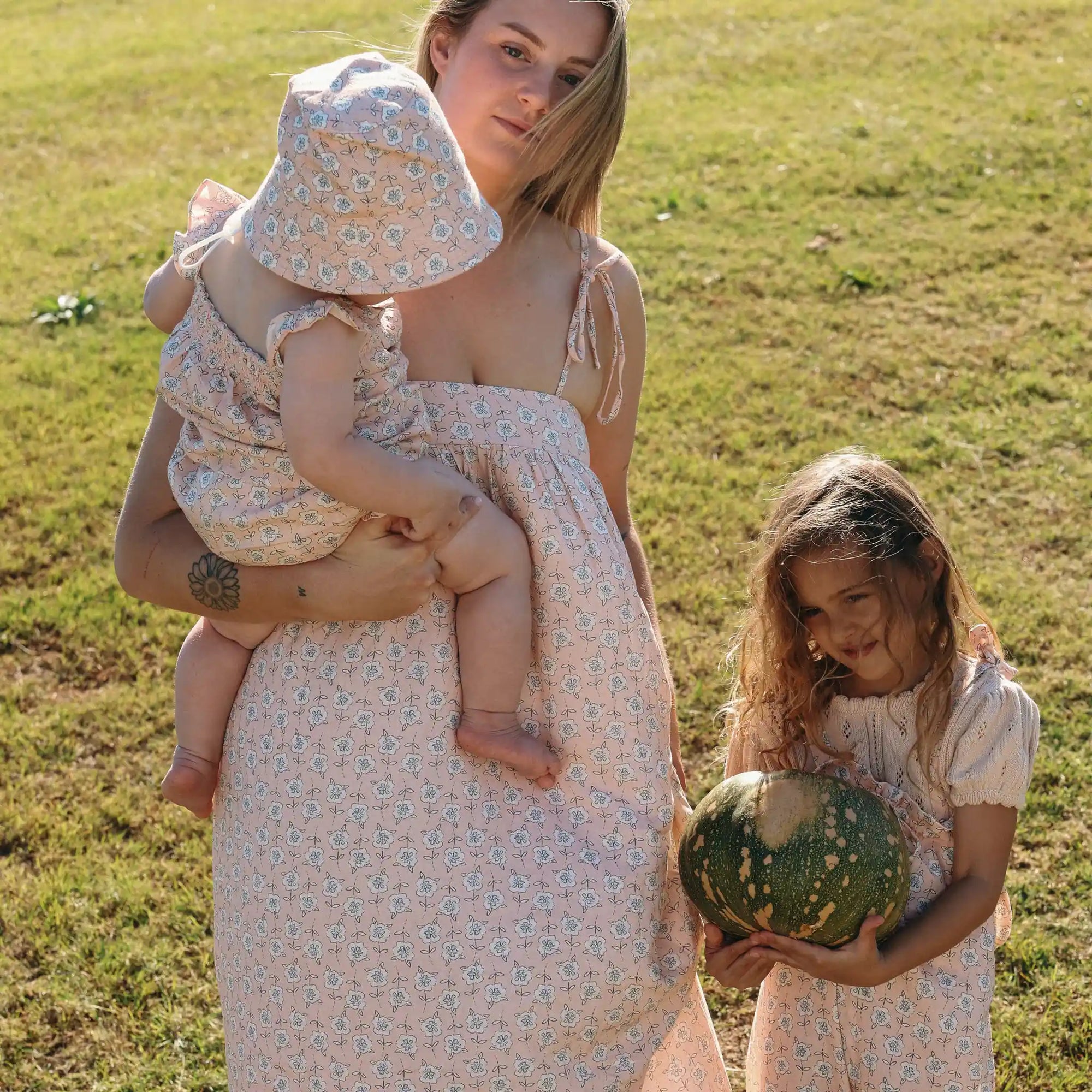 Mum in a floral print maxi dress holding a baby in a matching romper beside a girl carrying a pumpkin