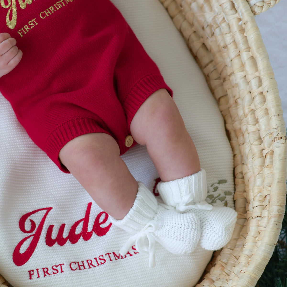 Close-up of a baby's legs in a red 'Jude's First Christmas' knit romper and white knit booties, lying on a matching personalised knit blanket in a woven bassinet