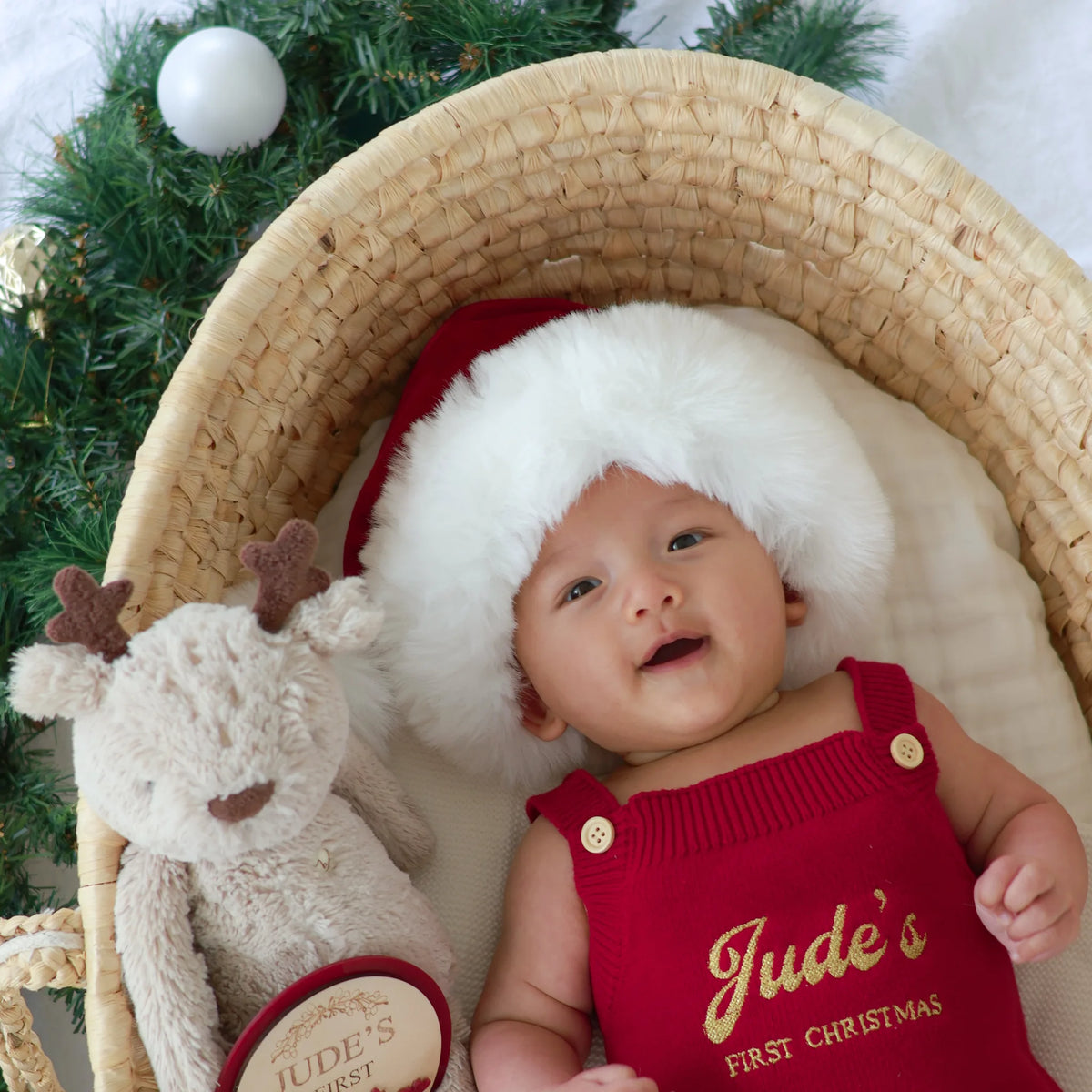 A baby wearing a red knit Christmas outfit with gold lettering and a white faux-fur Santa hat, lying in a woven basket next to a plush reindeer toy. The outfit reads 'Jude's First Christmas'.