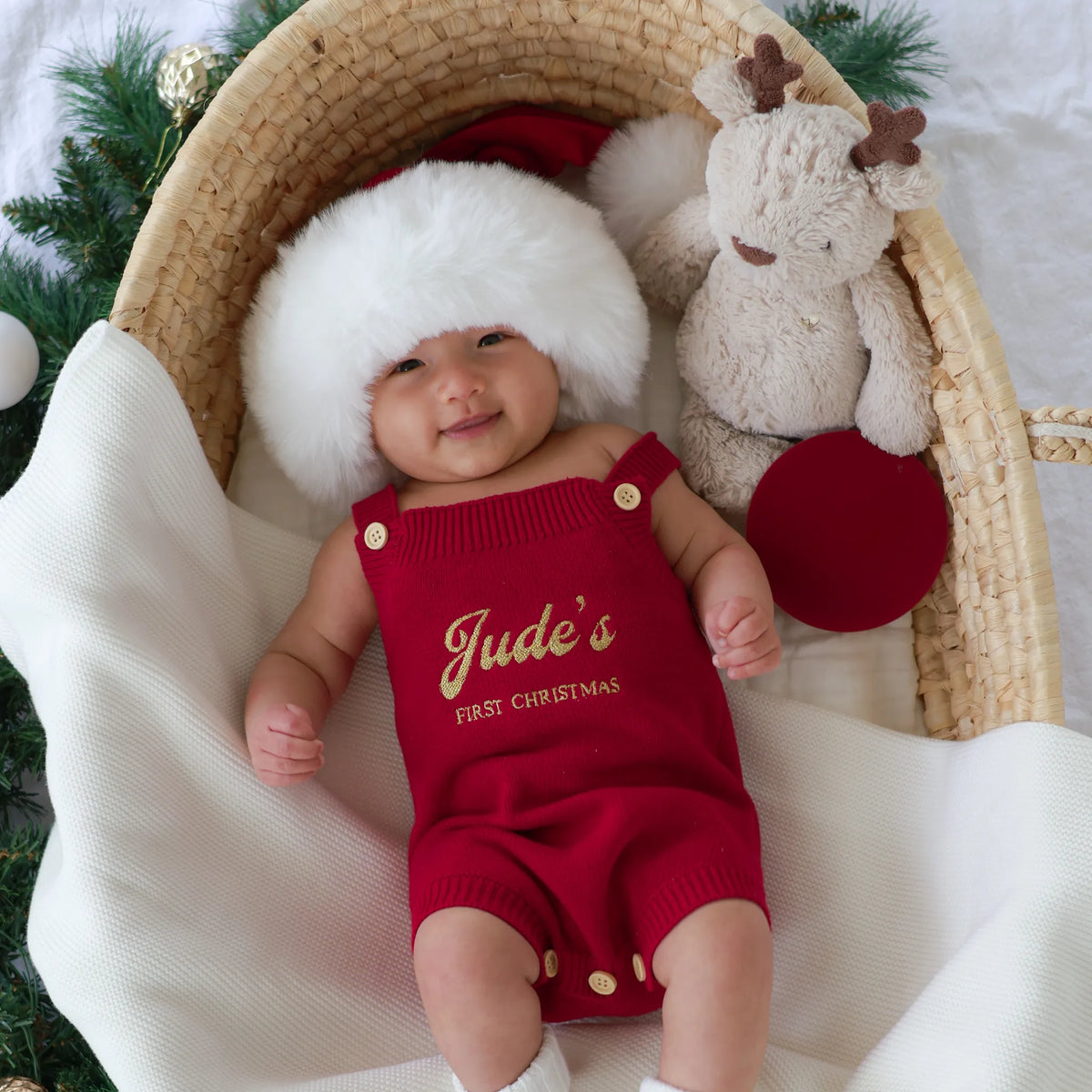A baby wearing a red knit romper with gold buttons and the words 'Jude's FIRST CHRISTMAS' in gold, styled in a wicker basket with a white faux-fur Santa hat, a plush reindeer toy, and a white blanket.