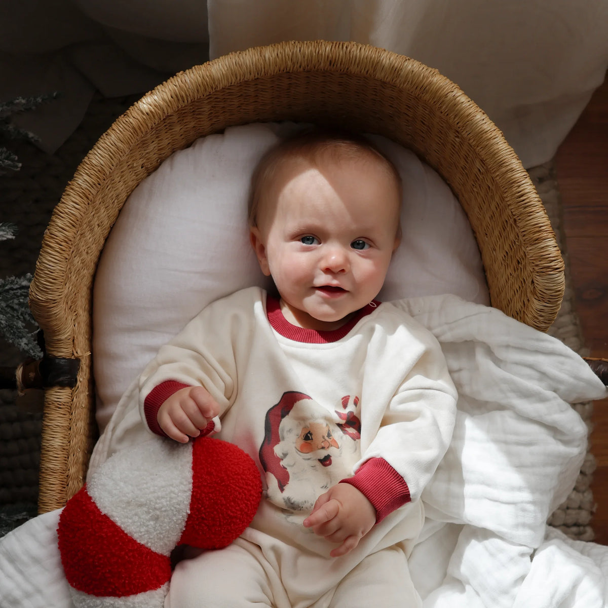 A baby lying in a wicker bassinet, wearing a cream-colored Christmas-themed footed sleeper with red cuffs and a Santa graphic, holding a red and white striped plush toy.