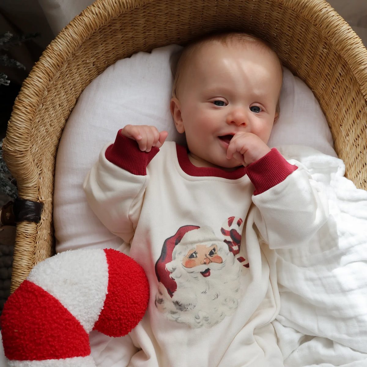 A baby wearing a white long-sleeve onesie with a Santa Claus graphic and red cuffs, lying in a woven wicker bassinet with a white cushion.