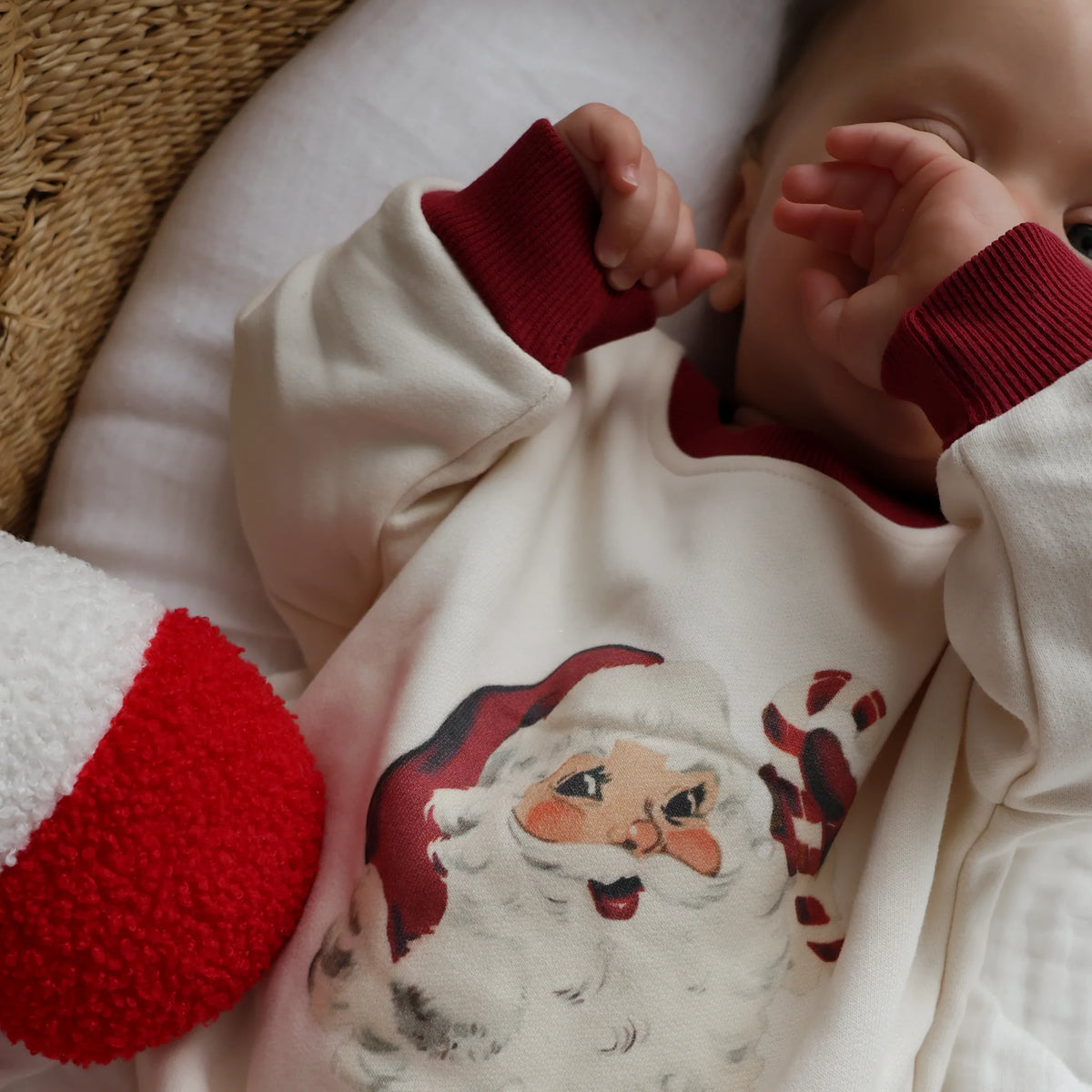 A baby wearing a cream-colored long-sleeve sweatshirt with dark red ribbed cuffs and a printed Santa Claus face graphic on the front, lying on a white cushion next to a red and white plush toy.