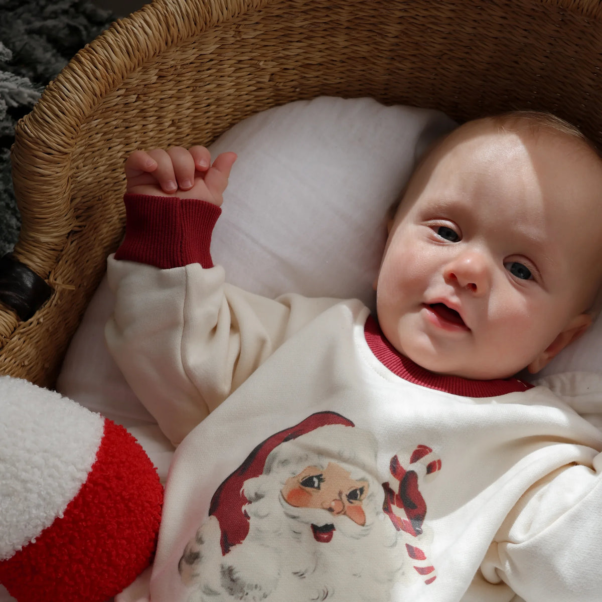 A baby lying in a wicker basket, wearing a cream long-sleeve shirt with a Santa Claus graphic and red ribbed collar and cuffs.