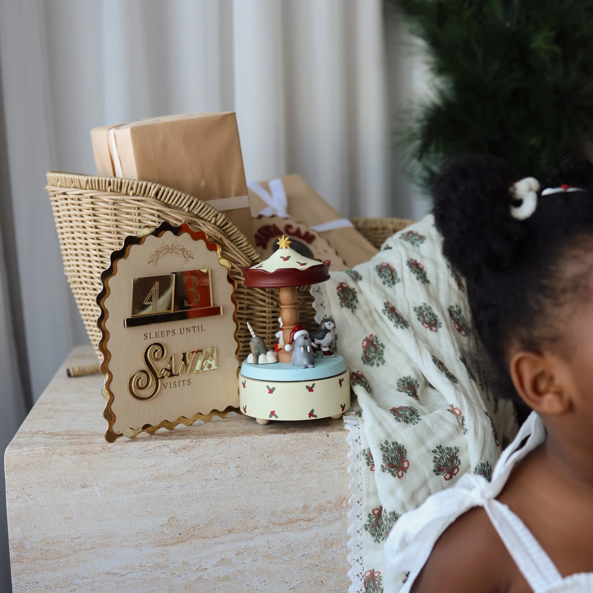 A styled holiday gift scene featuring a wicker sled with kraft paper wrapping, a decorative sign that reads 'SLEEP UNTIL SANTA', a small carousel music box, and a patterned cream blanket, with a child partially visible on the right.