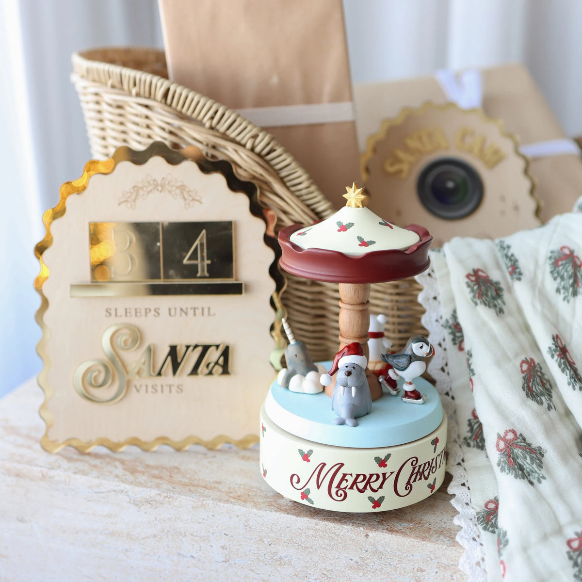 A festive Christmas-themed music box with penguins on a carousel, styled alongside a wooden basket, a gold-toned Santa visitor clock, and a patterned fabric on a light wooden surface.
