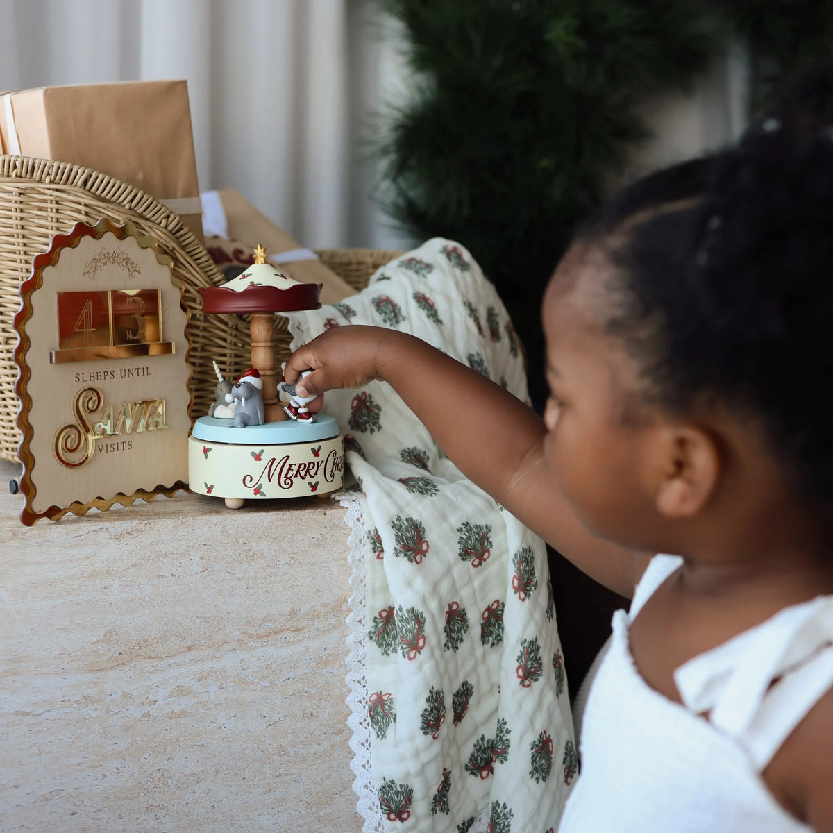 A young child in a white top reaches toward a small holiday-themed music box on a table. The box has a Santa-themed top and a cream base with 'Merry Christmas' written on it.