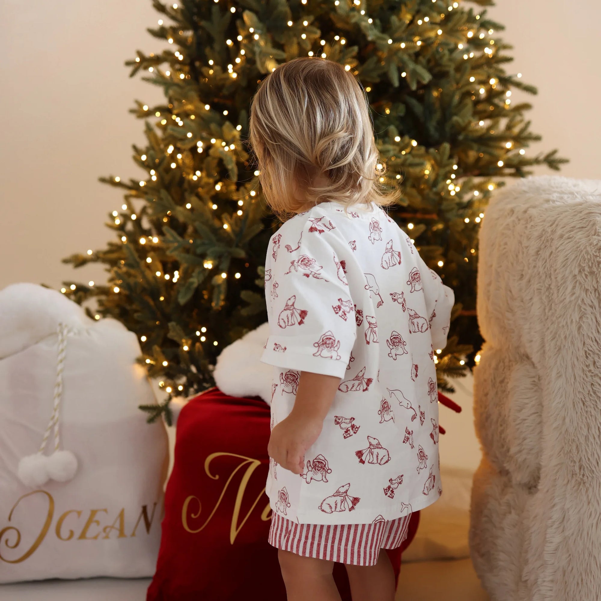 Back view of a toddler in a Christmas bear print shirt and red stripe shorts beside personalised Santa sacks