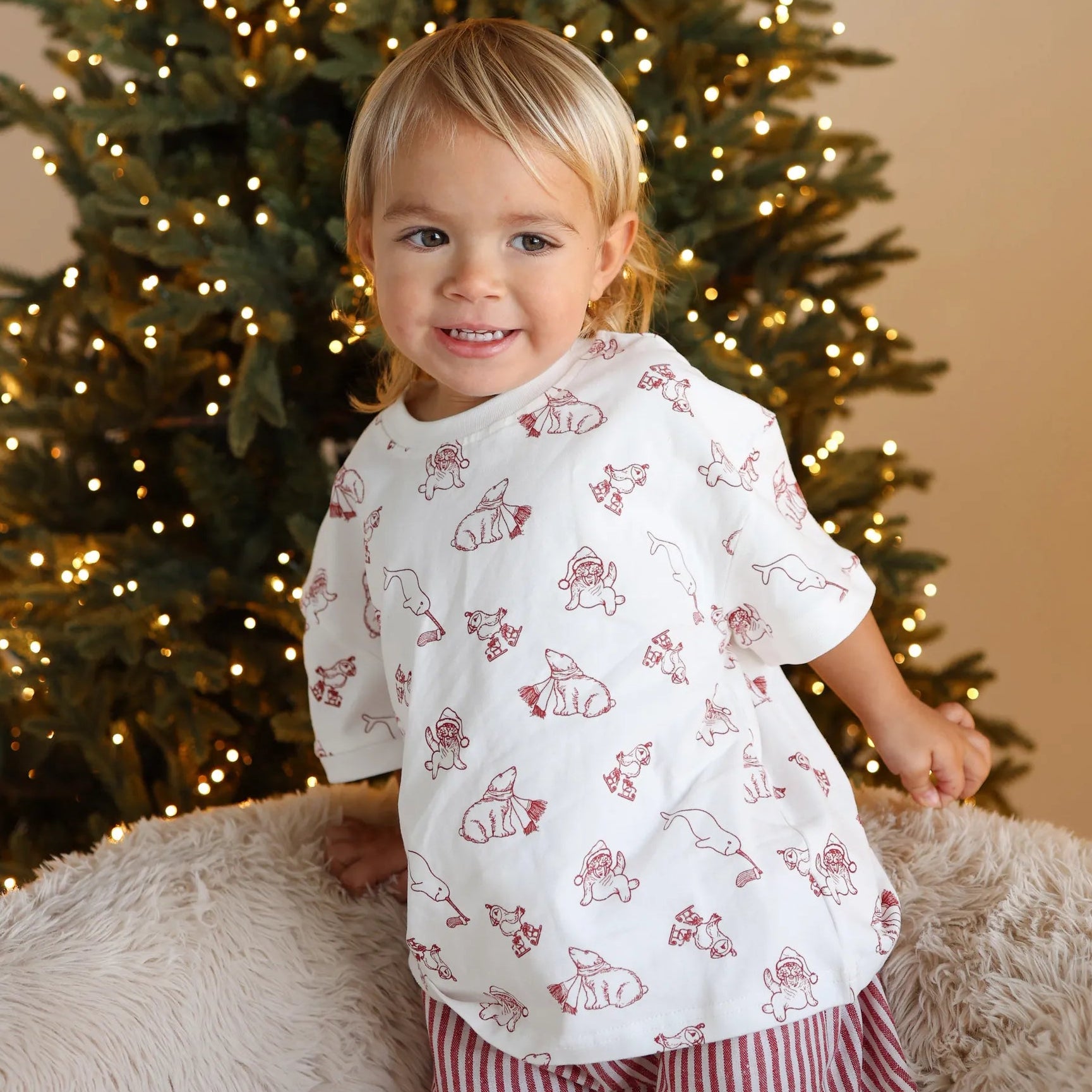 Toddler in a Christmas bear print shirt and red stripe shorts smiling in front of a lit Christmas tree