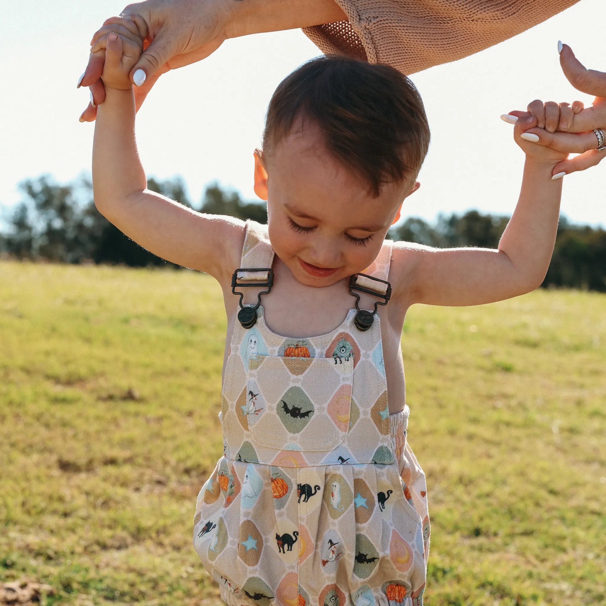 Toddler taking first steps in a Halloween print overall romper, held by both hands in a sunlit field