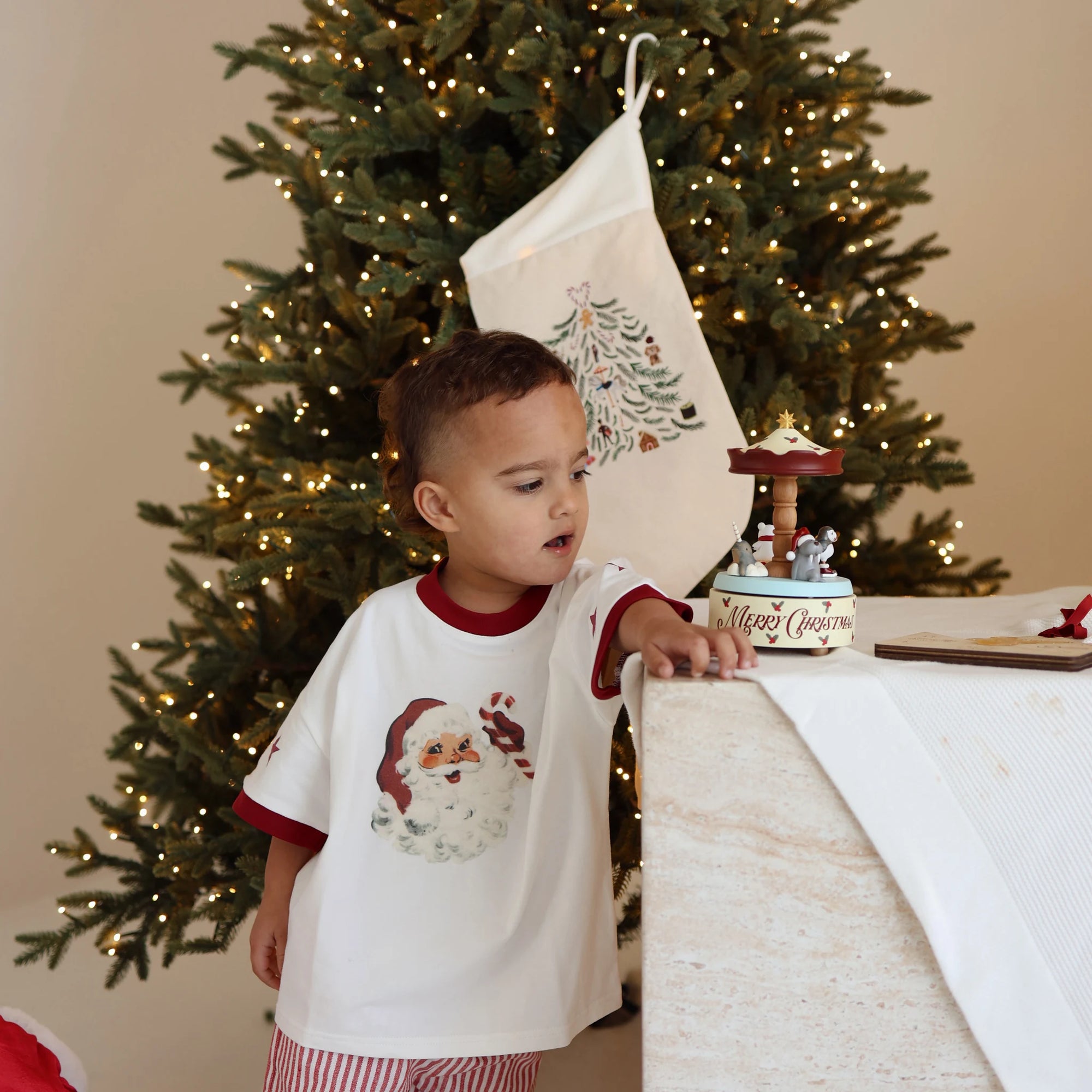 Toddler in a Santa print tee and red stripe shorts watching a Christmas carousel music box turn