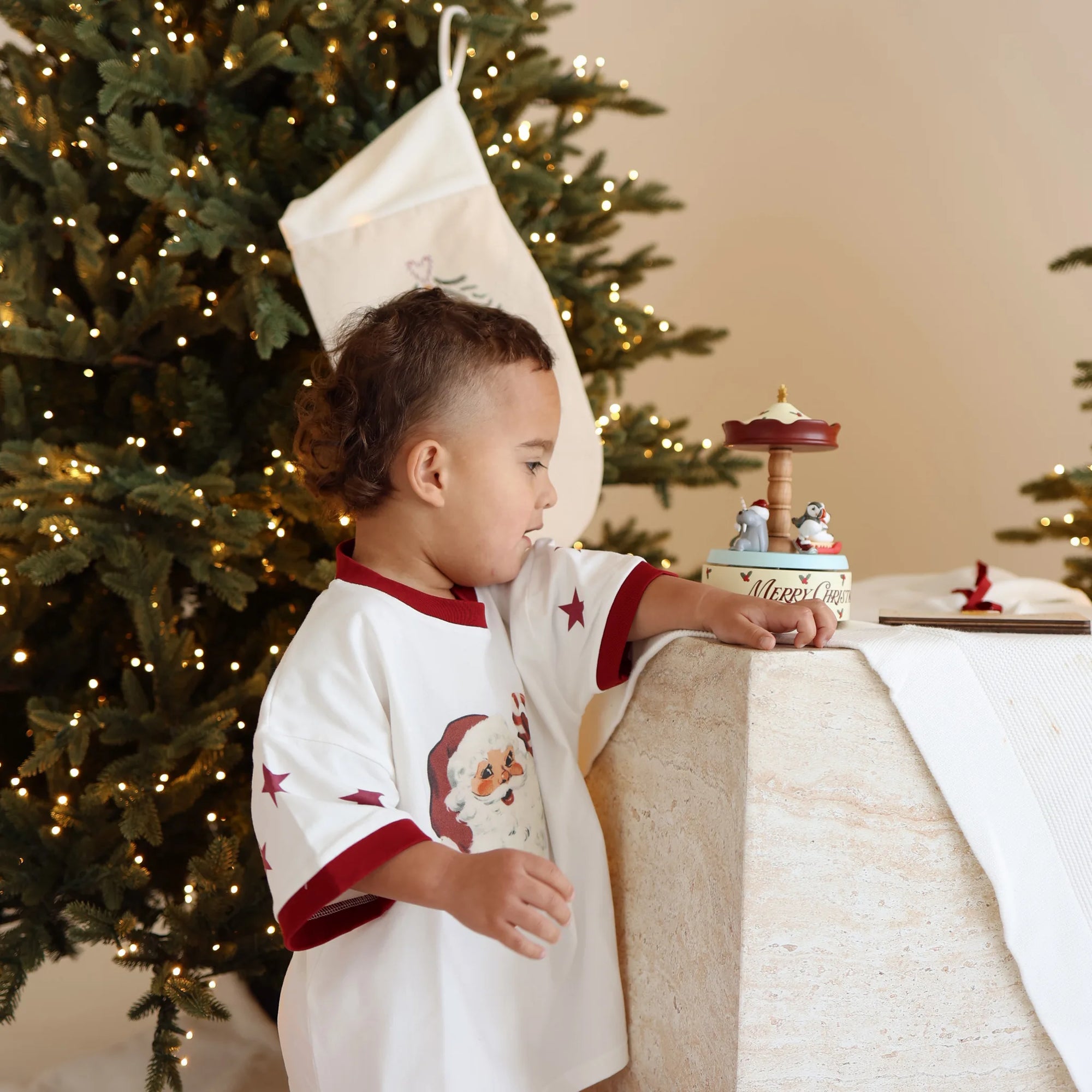 Toddler in a Santa print tee reaching for a Merry Christmas carousel music box beside an embroidered stocking